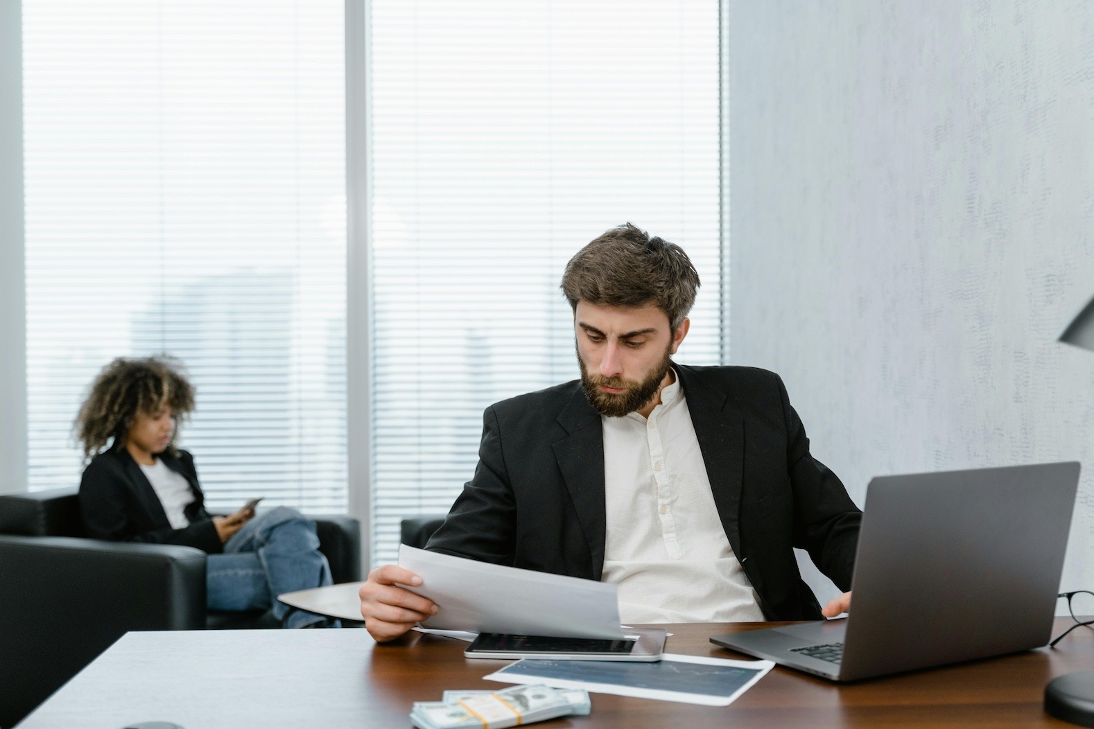 A man wearing a black suit jacket sitting at his office desk holding a piece of paper he is scanning. He is sitting in front of his open laptop.