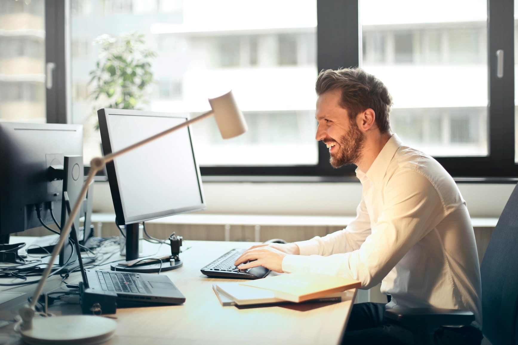A man working in an office looking at his computer monitor smiling. He is wearing a button up white collared shirt and has brown hair and beard.